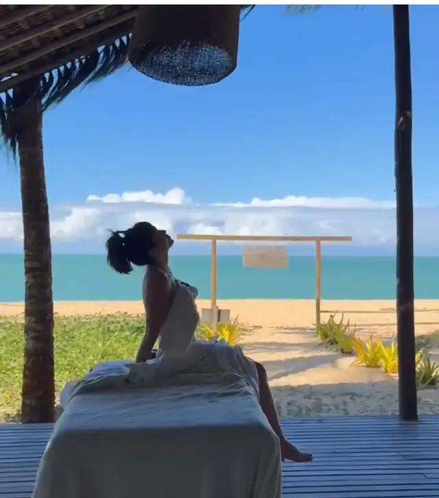 Mulher sentada na maca de massagem sob a cobertura de palha, com a praia e o mar azul ao fundo